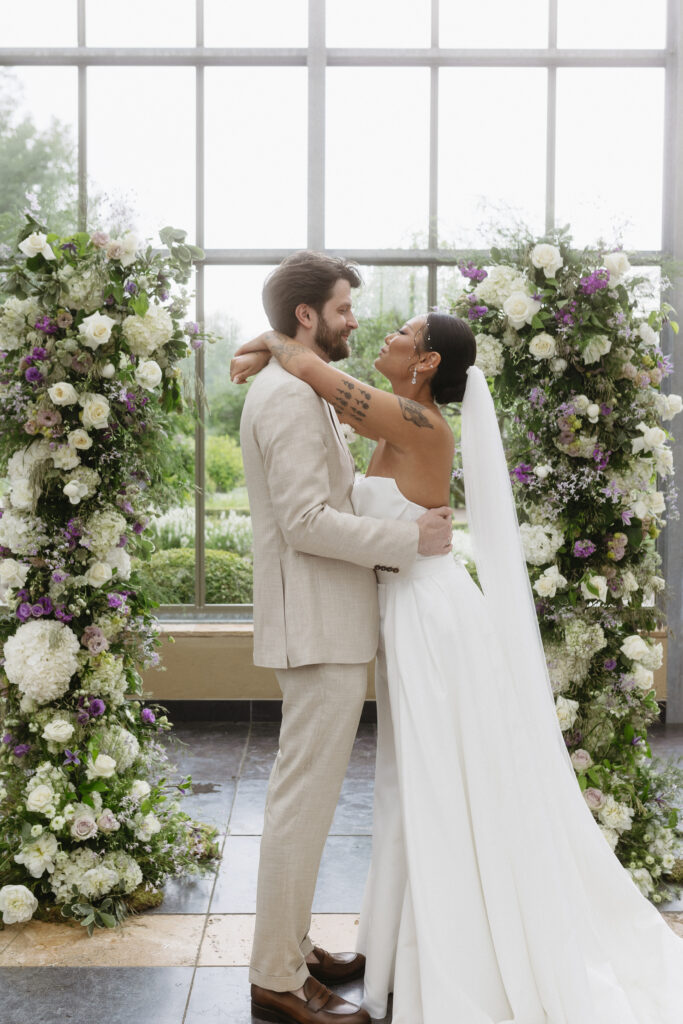 Modernes Hochzeitspaar dass vor dem Traubogen in einer Orangerie sich umarmen.